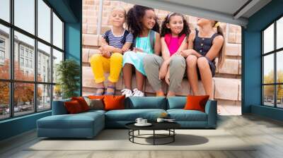 Full length of multiracial smiling elementary schoolgirls sitting on steps against school building Wall mural