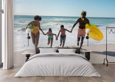 Happy african american family holding hands while running together at beach against clear sky Wall mural