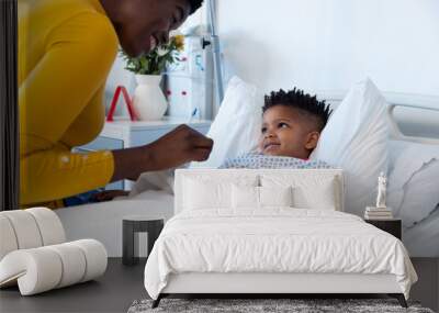 Happy african american mother sitting with son patient in hospital bed, smiling with copy space Wall mural