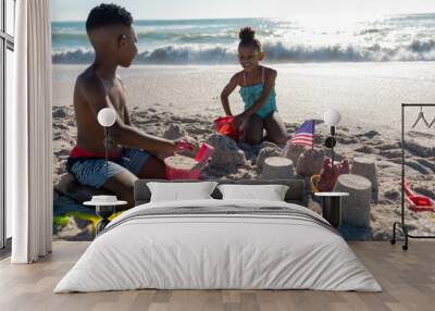 Happy african american siblings making sandcastle together at beach on sunny day Wall mural