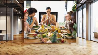 Happy diverse friends sitting at table and praying before dinner at christmas Wall mural