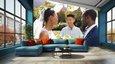 Happy diverse male officiant with bride and groom at outdoor wedding ceremony in sunny garden Wall mural