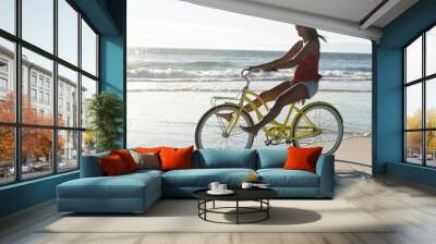 Happy mixed race woman on beach holiday riding bicycle on the sand by the sea with feet up Wall mural