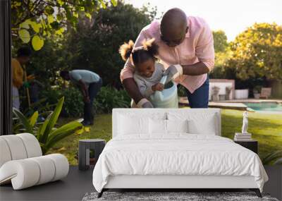 Happy senior african american man and his granddaughter watering plants in sunny garden Wall mural