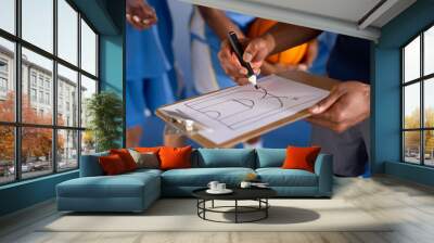 Male coach drawing basketball play on clipboard, players listening attentively in gym Wall mural