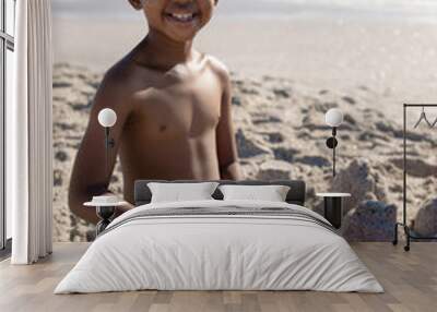 Portrait of smiling shirtless african american boy playing with sand pail and shovel at beach Wall mural