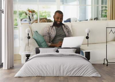 Smiling african american man using laptop and sitting on couch in living room Wall mural