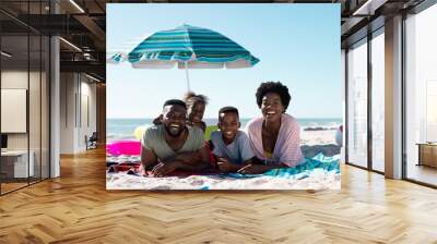 Smiling african american parents and children lying on towels at sandy beach under clear sky Wall mural