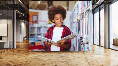 Smiling african american schoolboy reading book standing in school library Wall mural