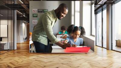 Smiling african american young male teacher assisting african american schoolgirl at desk in class Wall mural