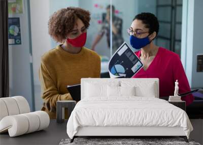 Two mixed race women wearing masks talking in an office Wall mural