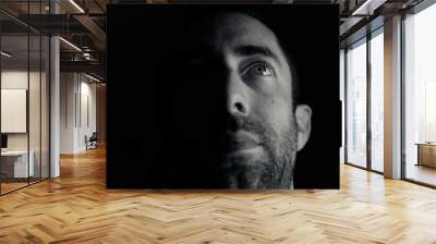 Dramatic portrait of a young man half expressionless face in deep shadow looking up isolated on black background Wall mural