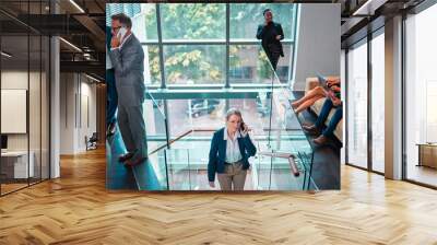 Arriving at work for the day. Shot of a mature businesswoman walking up the stairs while using a phone in a busy office at work. Wall mural