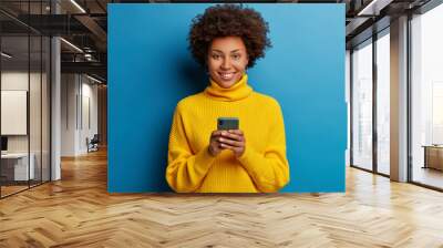 Adorable dark skinned adult woman dressed in yellow jumper, holds mobile phone, browses social networks, has broad smile, enjoys online chatting, isolated on blue wall, being always in touch Wall mural