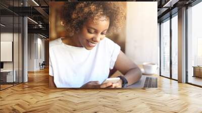 Adorable hipster dark-skinned woman with Afro hairstyle checking her news feed or messaging via social networks, using free wi-fi on mobile phone, smiling, sitting at cafe in front of laptop computer Wall mural