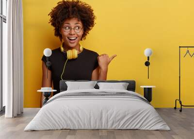 African American woman in high spirit points away with thumb, feels entertained and satisfied, listens radio broadcasting with modern headphones connected to telephone, checks sound, shows free space Wall mural