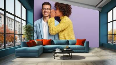 Afro American female whispers secret to boyfriend who has cheerful expression, gossip together, wear casual clothes, stands against purple background with blank space. Diverse couple indoor. Wall mural