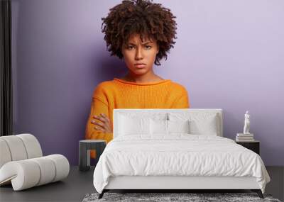 Angry sad young black female model crosses hands over chest, looks with grumpy expression, wears orange jumper, has Afro haircut, isolated over purple background. Negative human emotions concept Wall mural