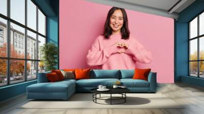 Attractive brunette young Asian woman feels happy and romantic shapes heart gesture expresses tender feelings wears casual jumper poses against pink background. People affection and care concept Wall mural