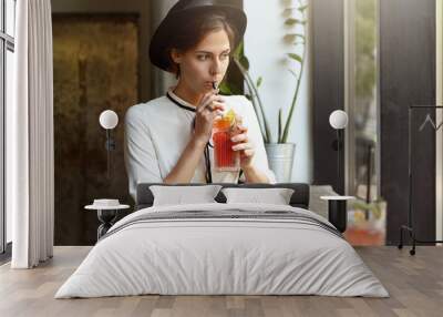 Attractive cute Caucasian girl in white jeans, blouse and black headwear sipping cold citrus juice or lemonade, sitting at cafe table by the window alone, looking outside with thoughtful expression Wall mural