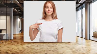 Attractive female in casual white t shirt, indicates at copy space for your logo, isolated over studio wall. Smiling teenage girl advertises new outfit, stands indoor. People, design, clothing concept Wall mural