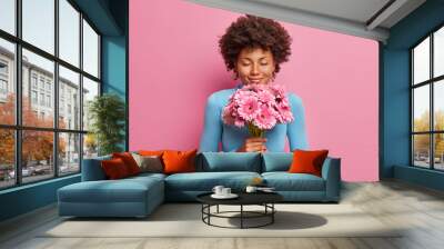 Attractive pleased dark skinned model receives flowers as present, stands with closed eyes, enjoys her favorite gerberas, isolated on pink background. Romantic portrait. Woman gets gift from husband Wall mural