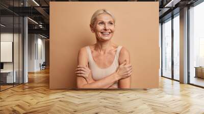 Attractive positive wrinkled fifty years old woman looks gladfully above keeps arms foded has well cared complexion healthy skin white teeth isolated over brown background. Beauty and age concept Wall mural