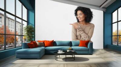 Attractive smiling young female customer with curly hair looking sideways and pointing her index finger at copy space on white blank studio wall for your text or promotional content. Horizontal Wall mural