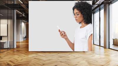 Beautiful African American female stands in profile, has serious look as reads text message on smart phone, connceted to wifi, surfes internet via modern device, isolated on white concrete wall Wall mural