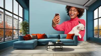 Beautiful cheerful dark skinned woman shoots online video about breed of dog for her blog, gives advice for followers how care this breed, takes picture of herself with pet via cellular, laughs Wall mural