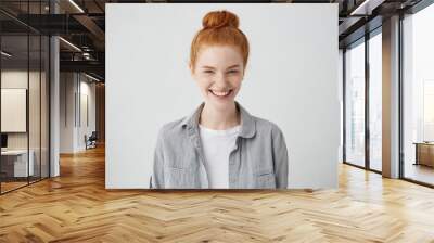 Beautiful ginger freckled girl with mysterious smile posing indoors at blank grey studio wall. Pretty woman with hair bun smiling broadly, showing white straight teeth, enjoying leisure time at home Wall mural