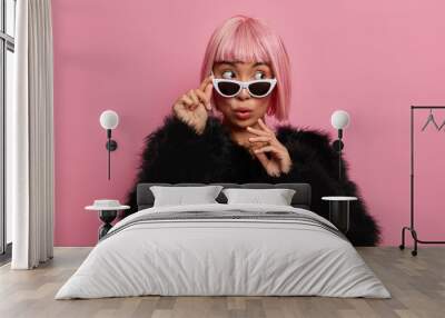 Beautiful wondered millennial girl wears pink hair wig, sunglasses and black fluffy sweater, scared to see something surprising and thrilling aside stands indoor over rosy wall. People, style, fashion Wall mural