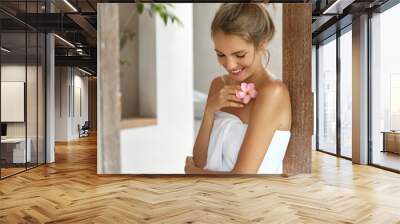 Beautiful young woman with soft skin, has light hair tied in knot, wears white towel after taking douche, smells flower, looks down with happy expression, pleased after good relax at bathroom Wall mural