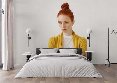 Beauty, people, youth and lifestyle concept. Studio shot of charming young redhead woman with freckles and hair knot staring at camera with serious look, crossing arms on her chest, dressed elegantly Wall mural