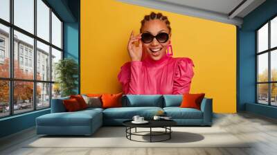 Beauty fashion model keeps hand on sunglasses, smiles positively, dressed in pink stylish clothes, being in good mood, rejoices buying new outfit, poses against yellow background. People and style Wall mural