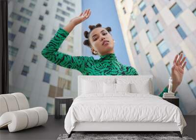 Below view of serious stylish teenage girl with makeup keeps arms raised dressed in comfortable clothes poses against modern skyscrapers against blue sky stands at downtown. Urban lifestyle concept Wall mural