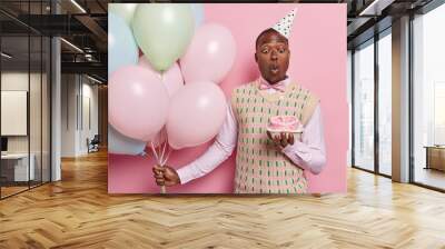 Birthday party. Indoor photo of African american man standing isolated on pink background wearing festive clothes holding colourful balloons blowing out candle on cake waiting for good presents Wall mural