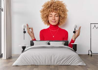 calm concentrated woman with curly hair keeps eyes closed and meditates indoor makes mudra gesture w Wall mural