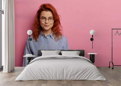 Calm optimistic red haired European woman looks directly at camera, has relaxed facial expression, charming smile, wears optical glasses and striped shirt, stands against pink background empty space. Wall mural