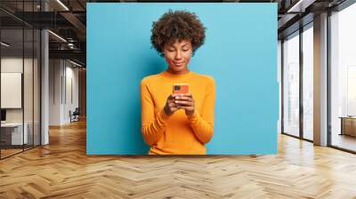 Calm serious dark skinned young woman plays games on phone or sends text messages connected to high speed internet uses modern technologies dressed in casual jumper poses against blue background. Wall mural