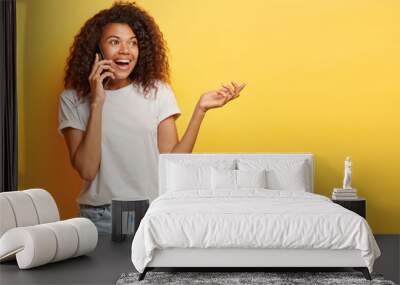 Candid shot of carefree optimistic girl with bushy Afro hairstyle talks with friend about future vacation, enjoys good roaming connection focused aside dressed in casual outfit isolated on yellow wall Wall mural