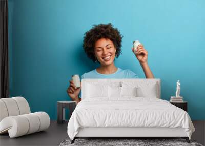 Carefree joyful dark skinned woman holds two jars of fresh organic yoghurt smiles joyfully keeps to healthy diet eats delicious food dressed in t shirt isolated on blue background. Dairy product Wall mural