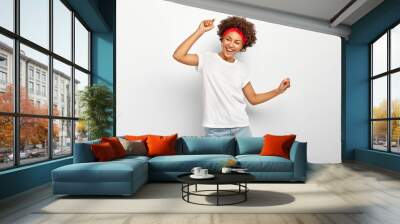 Carefree pleased teenage girl has fun, dances joyfully with raised arms, being entertained and amused, wears summer clothes, laughs happily, enjoys cool music, isolated on white wall, being on party Wall mural