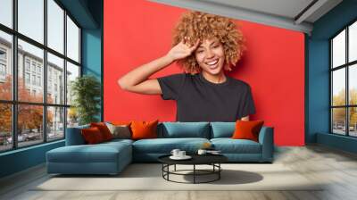 Carefree young woman smiles toothily keeps hand on forehead feels joyful wears casual black t shirt poses against vivid red background. Positive curly haired female model poses for making photo Wall mural