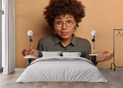 Careless hesitant woman shrugs shoulders, spreads arms sideways, cant help or has doubts, wears stylish shirt and big glasses, unable to give answer on confusing question, models in brown studio Wall mural