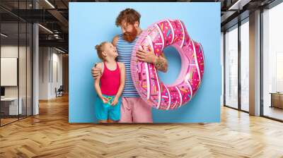 Caring father embraces little ginger daughter, holds inflated swimring, going to swim in sea, spend summer vacation together, have happy expressions, isolated on blue background. Family, vacation Wall mural