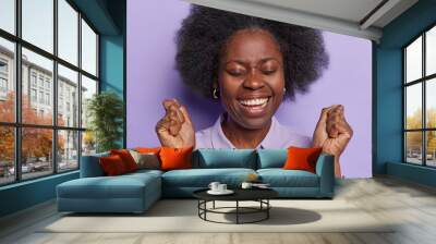 Charming optimistic African woman feels joyful smiles toothily shakesclenched fists keeps eyes closed and triumphs over something got success on work isolated on purple background. Yeah finally I won Wall mural