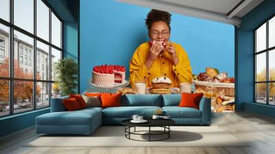 Cheat meal and gluttony concept. Ethnic curly woman eats strawberry creamy cake with much calories, has sweet lunch, tastes various desserts, leads sweet life with confectionery. Female fan of bakery Wall mural