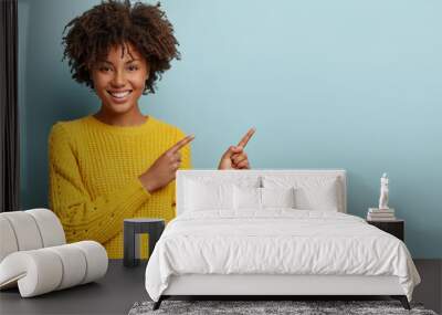 cheerful afro woman points away on copy space, discusses amazing promo, gives way or direction, wear Wall mural