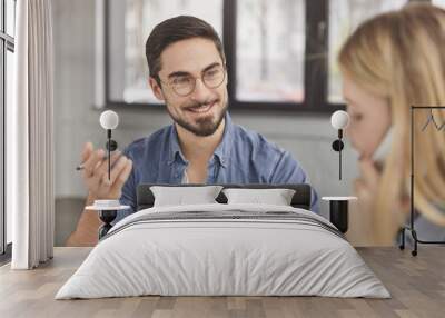 Cheerful bearded handsome male wears round glasses, has pleasant conversation with female colleague, try to discuss main issues of their work. Pretty young woman talks via modern cell phone. Wall mural